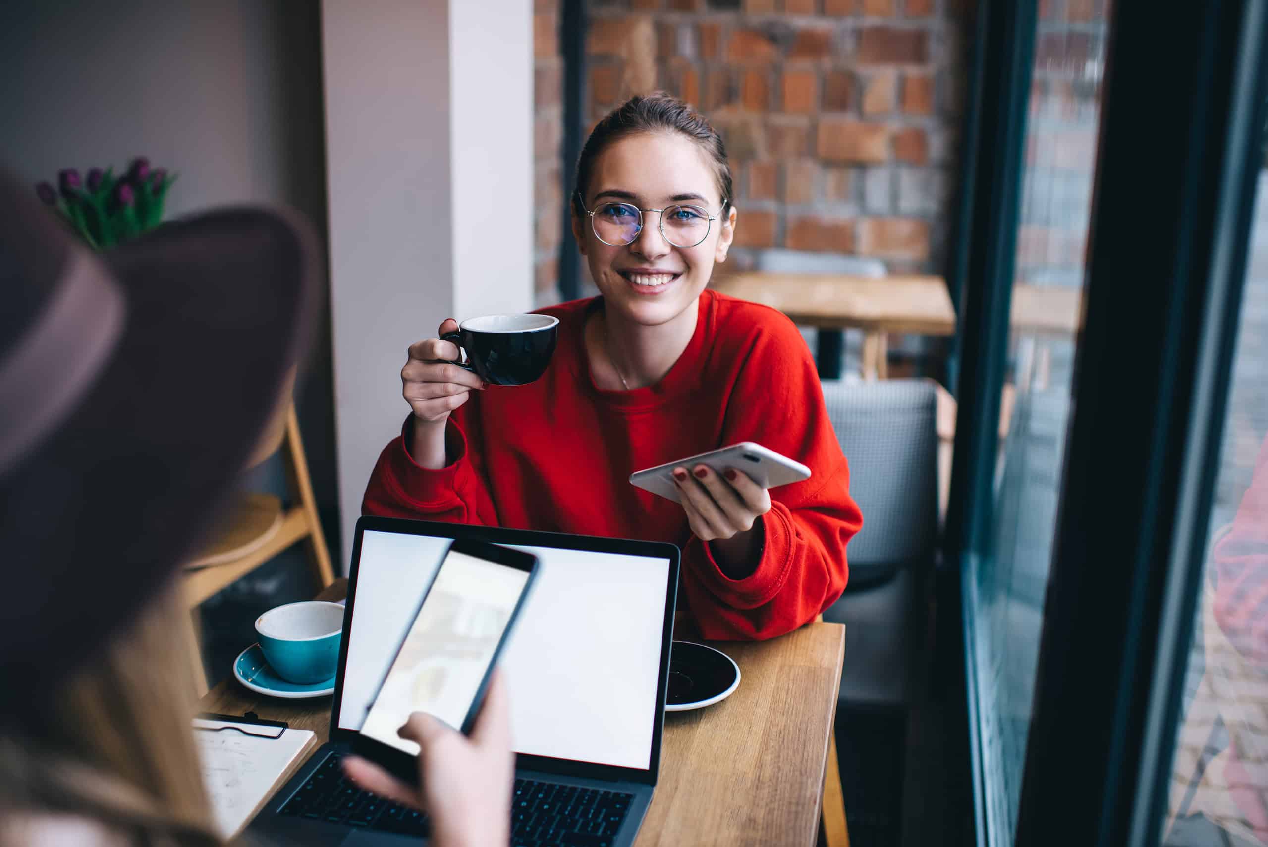 High angle of cheerful female in casual wear and glasses resting at wooden table with friend and messaging on smartphone while drinking coffee