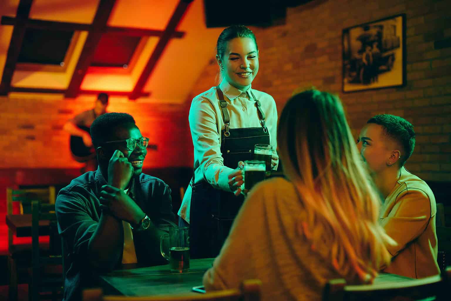 Group of happy friends enjoying while waitress is serving them beer in a pub. Focus is on waitress.