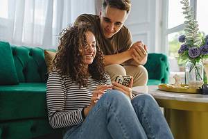 caucasian woman using mobile phone for online sms chat or internet browsing while her boyfriend or husband adult male sit beside at home in bright room real people couple leisure concept